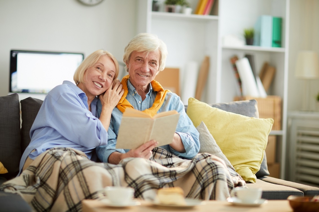 Portrait of loving senior couple looking at camera while reading book together sitting on comfortable sofa at home, copy space