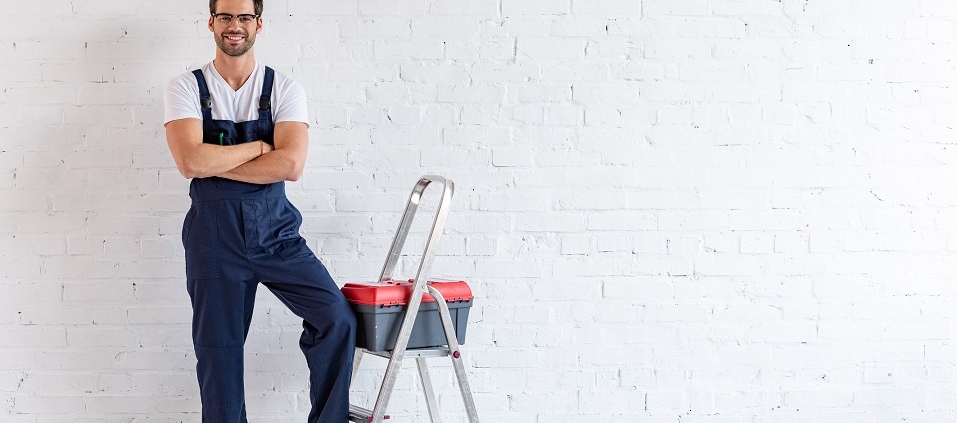 smiling repairman standing under air conditioner near stepladder and toolbox and looking at camera